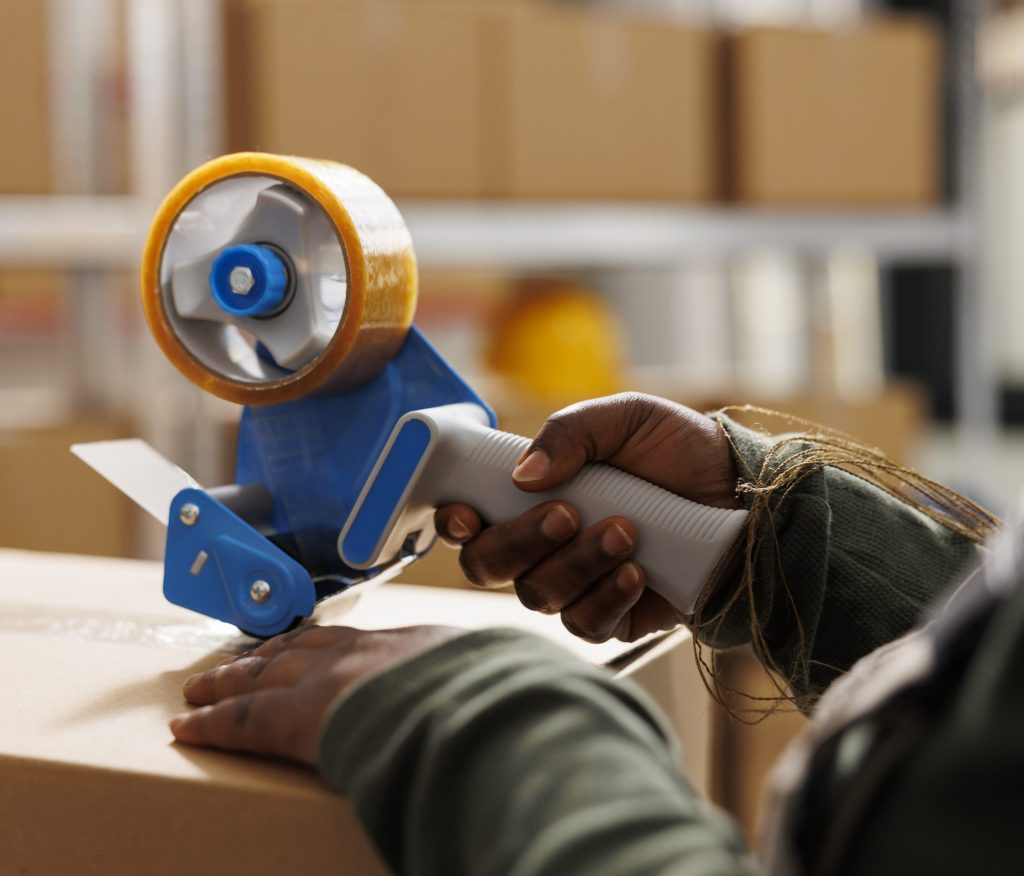 Storehouse worker using adhesive tape to pack clients orders, preparing packages for delivery in storehouse. African american employee wearing industrial overall during merchandise inventory. Close up